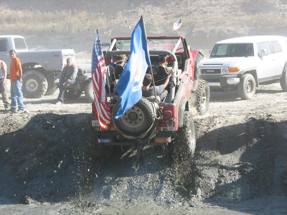 Red Jeep Wrangler with American and blue flags driving through mud, white Toyota FJ Cruiser and Ford truck in background