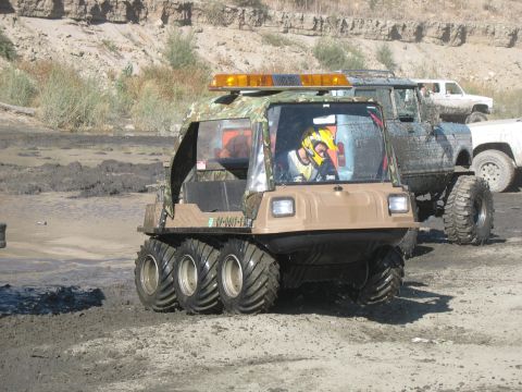 1987 Ford Bronco - An amphibious vehicle with six wheels navigates a muddy terrain, with other trucks visible in the background.