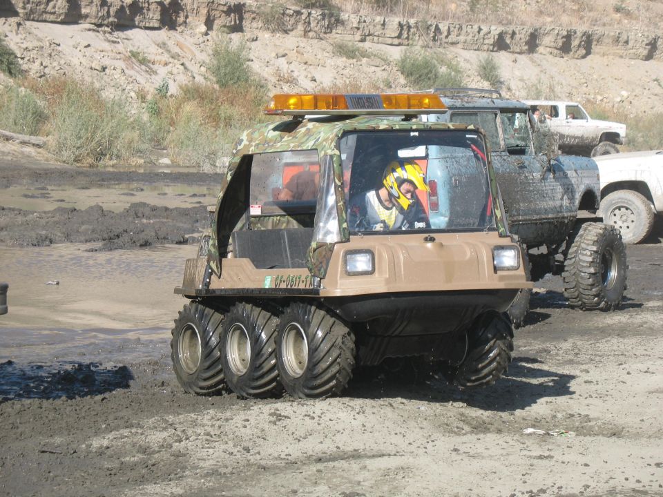 Tan 6x6 amphibious ATV with camo top and light bar driving through a mud pit