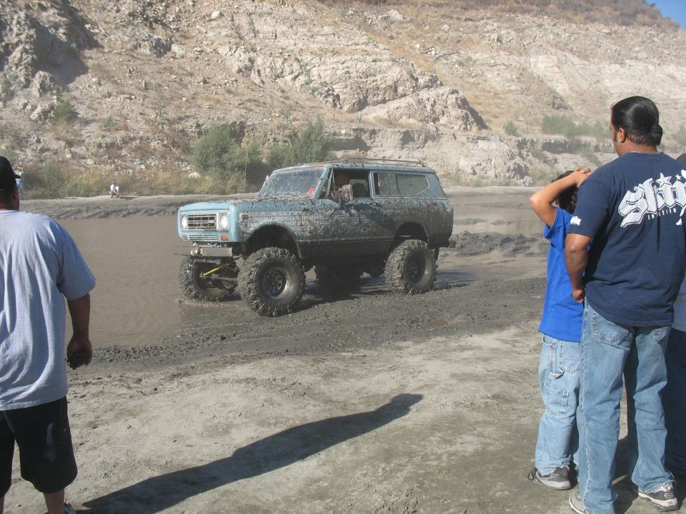 Mud-covered SUV with large tires driving through shallow water, people watching