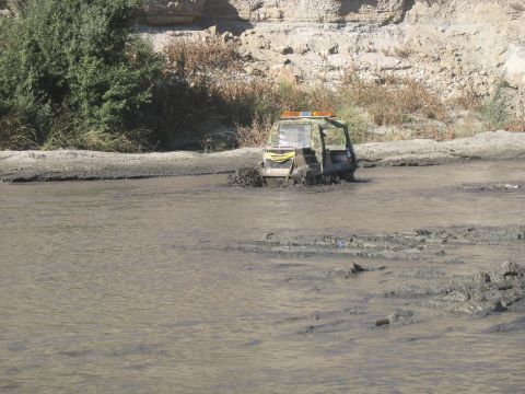 1987 Ford Bronco - An off-road vehicle navigates through a muddy, shallow body of water with vegetation and a rocky embankment in the background.