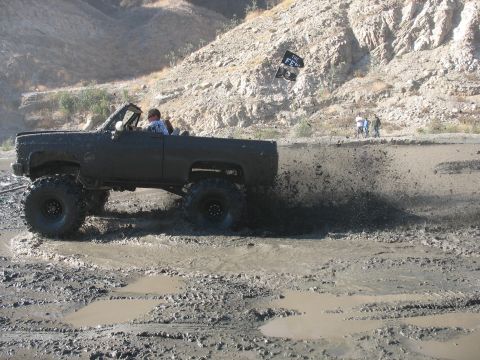 1987 Ford Bronco - Off-road pickup truck navigates a challenging muddy terrain, sending mud flying.