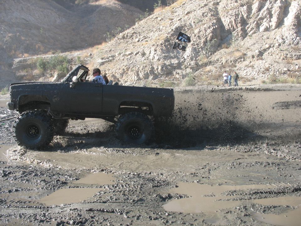 Lifted pickup truck with large tires driving through a muddy area, kicking up mud