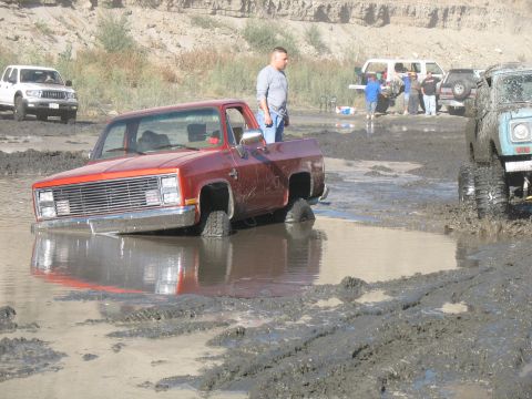 1987 Ford Bronco - A red pickup truck is stuck in a mud pit, with other vehicles and people in the background.