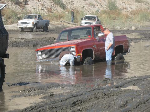 1987 Ford Bronco - A red pickup truck is stuck in a muddy waterhole with two people observing.