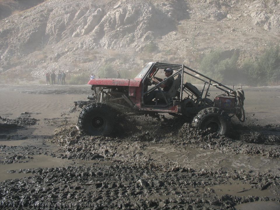 Red tube-cage rock buggy driving through deep gray mud with a rocky hillside behind