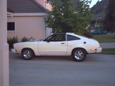 1978 Ford Mustang II - Full side profile of a white Mustang II hatchback parked on a concrete driveway at dusk, wearing a black roof panel, polished five-spoke ma…
