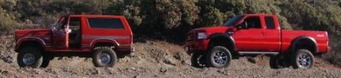 1986 Ford Bronco - A red 1986 Ford Bronco and a red pickup truck are parked side-by-side on a rocky, wooded hillside.