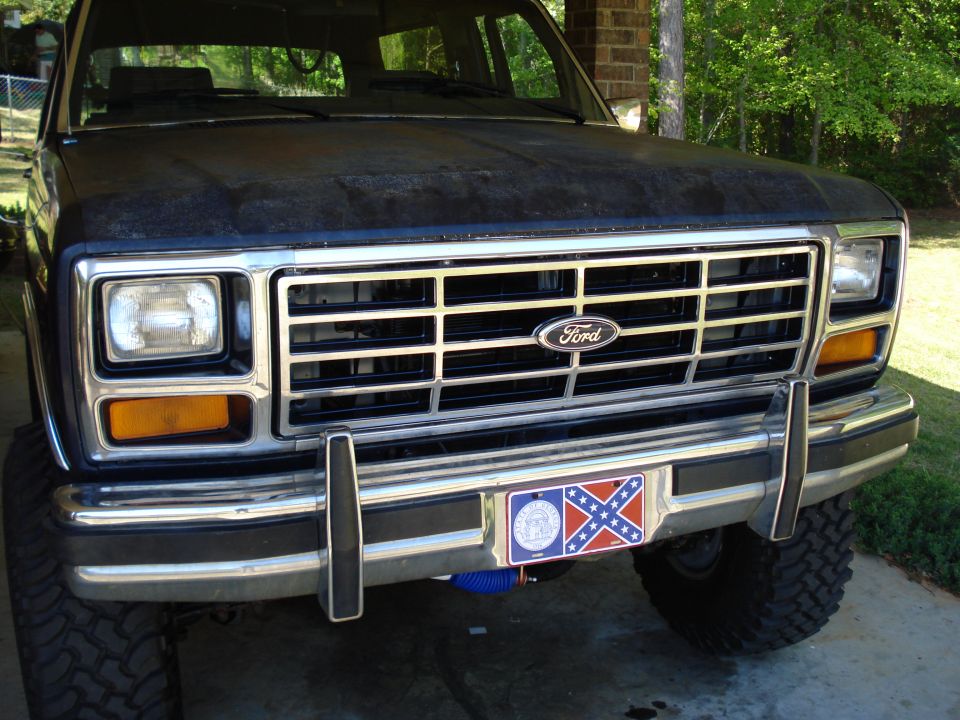 Front of a dark-colored Ford Bronco with a chrome grille and bumper. License plate has a Confederate flag design