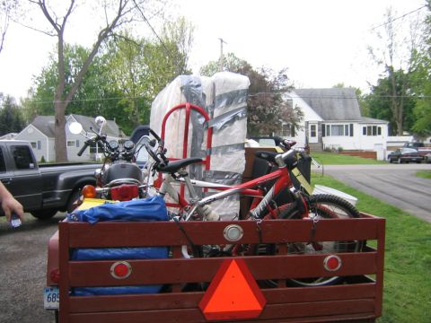 1994 Ford Bronco - A trailer carries a motorcycle, bicycles, and large wrapped items, with houses and trees in the background.