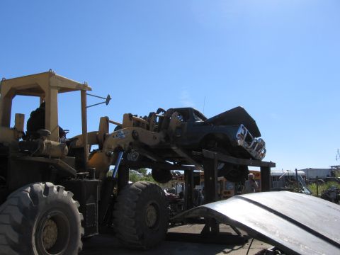 1995 Ford F-150 - A loader lifts the front end of a dark pickup truck, showing its grille and headlights.