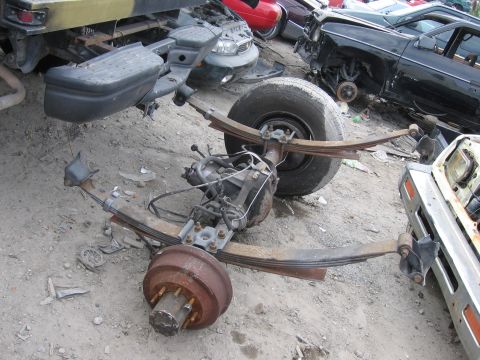 1995 Ford F-150 - A disassembled rear axle assembly sits on the ground in a junkyard, surrounded by other vehicle parts and wreckage.