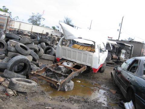 1995 Ford F-150 - A damaged pickup truck sits in a junkyard, surrounded by debris and other wrecked vehicles.