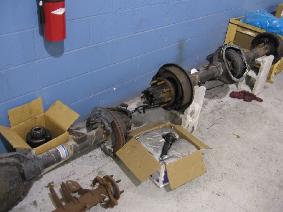 Two vehicle rear axles are on the floor, one in a box. A red fire extinguisher is on the wall