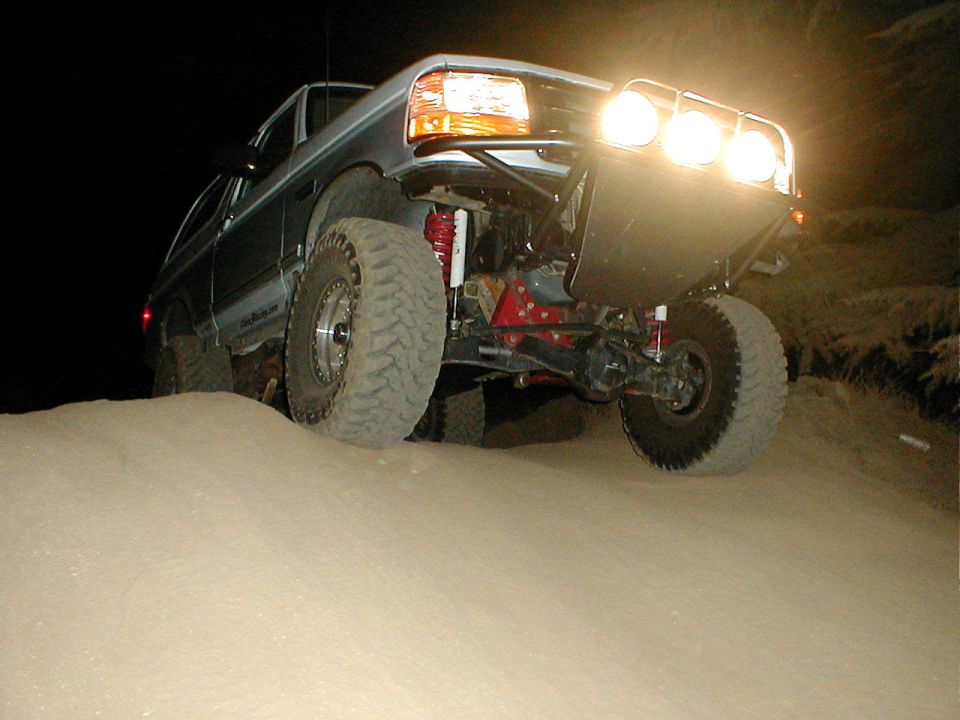 Silver Ford Bronco with off-road tires and lights, angled from the front right, driving on sand