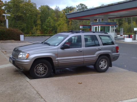 1991 Ford Crown Victoria - A silver 1999 Jeep Grand Cherokee is parked on a sidewalk near a gas station with a "Caisse" booth.