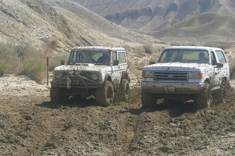 1990 Ford Bronco - Two SUVs navigate a muddy trail in a desert landscape, kicking up dirt as they go.