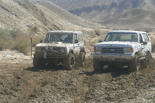 Two muddy SUVs, one older Bronco-style and one newer Ford, driving through a dirt track in a desert canyon
