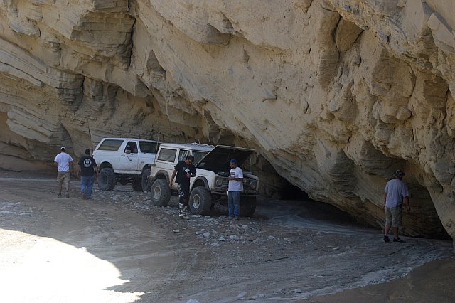 Two white SUVs parked on a dirt road under a large rock overhang. People are standing around them