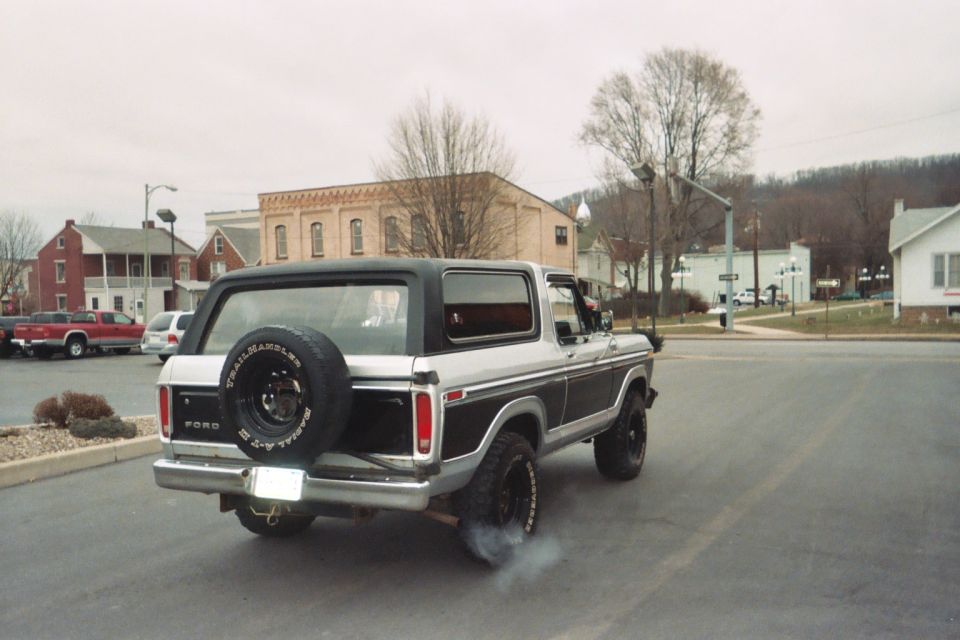 1978 Ford Bronco, two-tone silver and black, with spare tire mounted on the rear door, emitting exhaust smoke