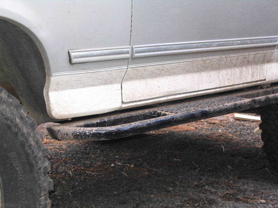 Muddy silver SUV side with black step bar and large off-road tires on gravel