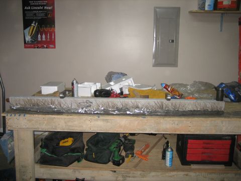1996 Ford Bronco - Cluttered home garage workbench topped with a long welded-and-taped fabrication piece, aluminum boxes, spray cans, welding gloves and hand…