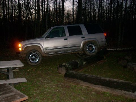 2000 Chevrolet Tahoe - A silver SUV navigates an obstacle course of logs in a wooded area at dusk.