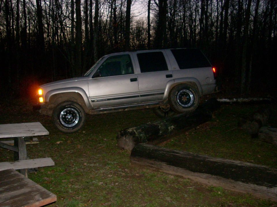Silver SUV on logs, front wheel elevated. Dark forest background. "SUPERMOTORS" text visible