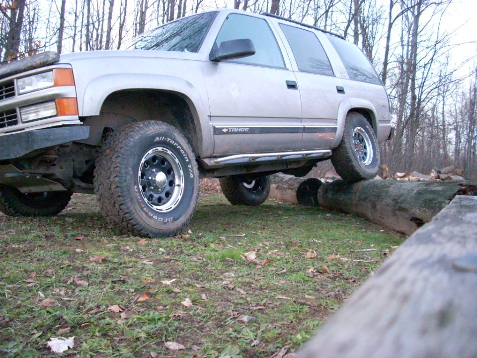 Silver SUV with off-road tires driving over logs. The vehicle appears to be a 1995 Chevrolet Tahoe