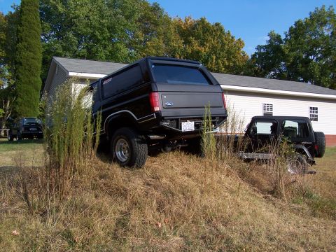 1983 Ford Bronco - Two off-road vehicles are parked on a grassy slope beside a building on a sunny day.