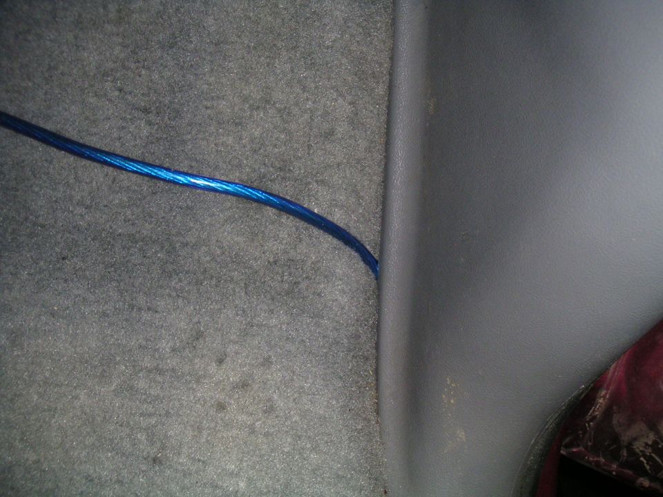 Blue wire running along gray carpet next to dark gray plastic trim inside a vehicle