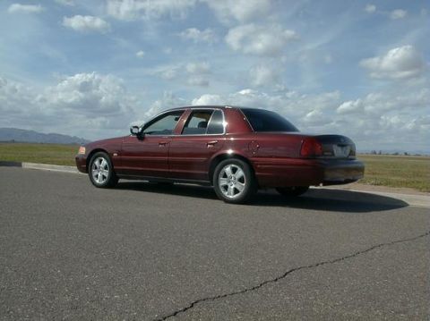 2004 Ford Crown Victoria - This maroon 2004 Ford Crown Victoria sedan is captured from a rear three-quarter angle, parked alongside a road under a cloudy sky.