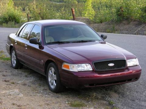 2004 Ford Crown Victoria - A maroon 2004 Ford Crown Victoria is parked on the side of a road with trees and a fence in the background.