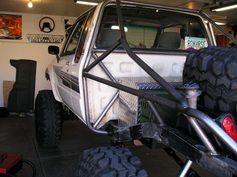 1986 Toyota Hilux - A white Toyota pickup mid-build shown from the front-left in a garage, fitted with a bare-metal tube roll cage extending over the cab and b…