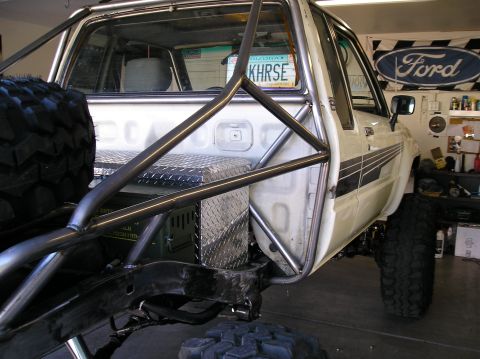 1986 Toyota Hilux - White lifted mini-truck in a garage, shot from the driver's side rear showing a bare-metal exo/roll cage tied into the bed, diamond-plate b…