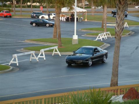 1995 Ford Probe - A black 1995 Ford Probe is captured in motion within a parking lot, with other vehicles and palm trees visible in the background.