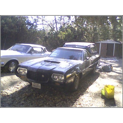 1972 Ford Ranchero - A dark 1972 Ford Ranchero with a camper shell is parked in a gravel lot next to a white car and a shed.