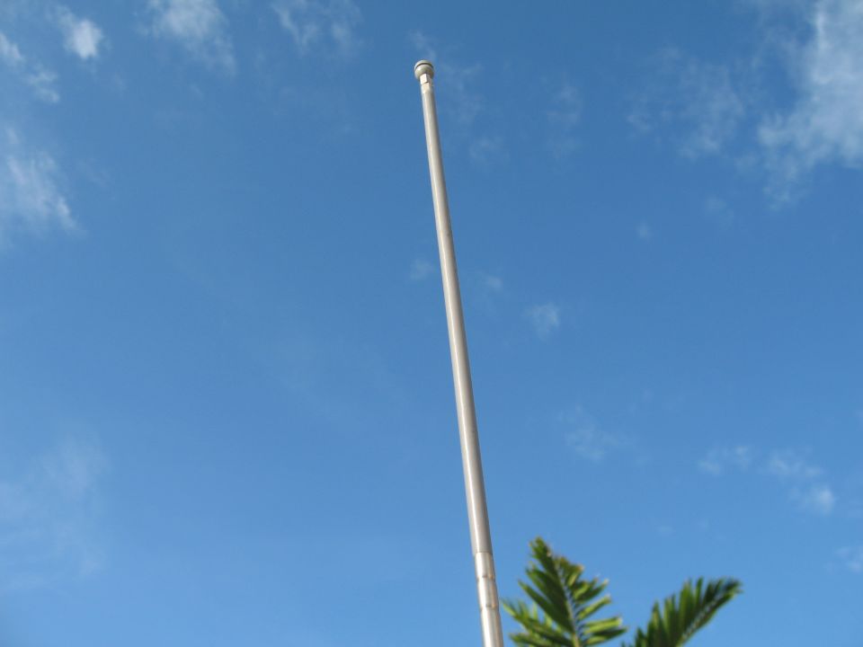 Tall CB antenna against a blue sky with clouds. Some green foliage is visible at the bottom right