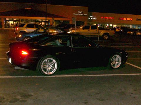 1995 Ford Probe - A black coupe is parked in a lot at night, with other vehicles and businesses in the background.