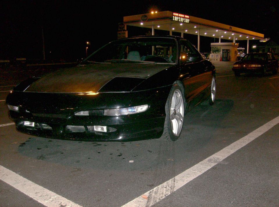 Black coupe with a matte hood parked at a gas station at night. Another car is visible in the background