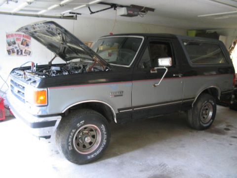 1989 Ford Bronco - A two-tone gray and black Ford Bronco is parked in a garage with its hood open, revealing the engine compartment.