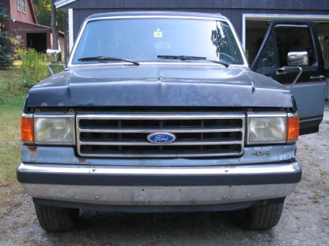1989 Ford Bronco - Front view of a gray and black Ford Bronco with the driver's door ajar, showing the grille and chrome bumper.