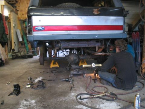 1989 Ford Bronco - A person works on the undercarriage of a lifted Ford Bronco in a garage setting.