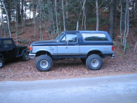 1989 Ford Bronco - A lifted Ford Bronco and a Jeep are parked side-by-side in a wooded area.