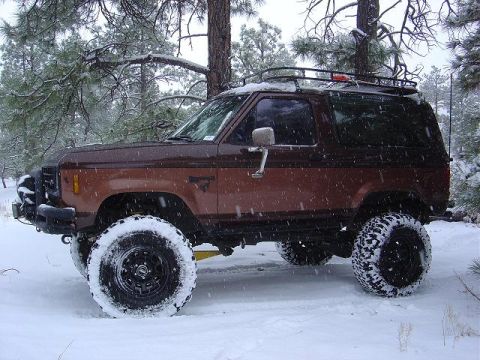 1956 Ford Bronco II - A brown Ford Bronco navigates a snowy landscape with its roof rack and large tires.