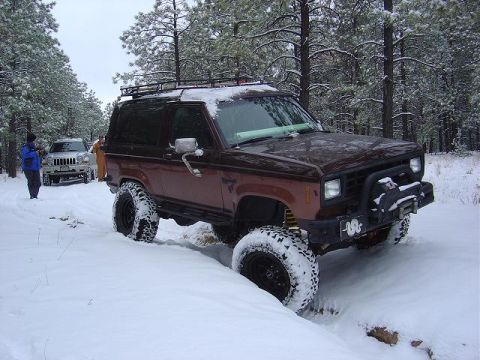 1956 Ford Bronco II - A lifted Ford Bronco II navigates a snow-covered forest road, with another vehicle visible in the background.