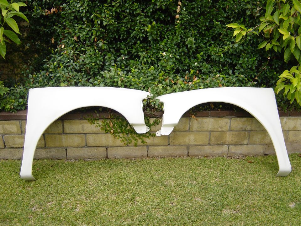Two white car fenders laid on grass in front of a brick wall and hedge