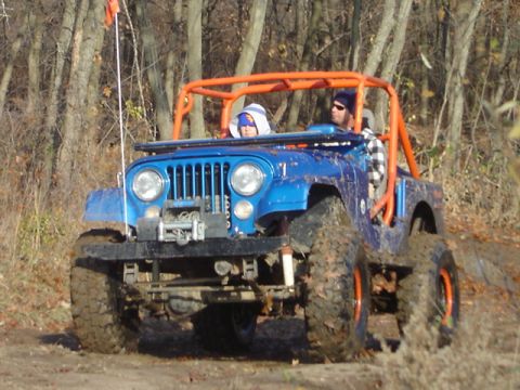 1992 Ford Bronco - A blue Jeep with an orange roll cage navigates a muddy trail with two occupants.