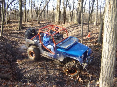 1992 Ford Bronco - A blue off-road jeep navigates a muddy, wooded path with an orange roll cage and two occupants.