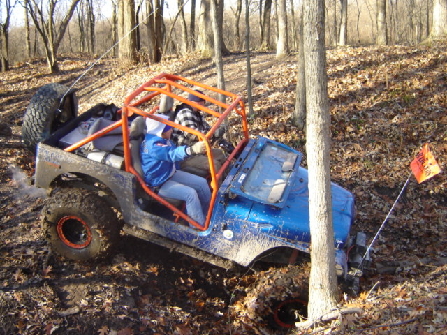 Blue off-road Jeep with orange roll cage stuck in mud, driver wearing blue jacket